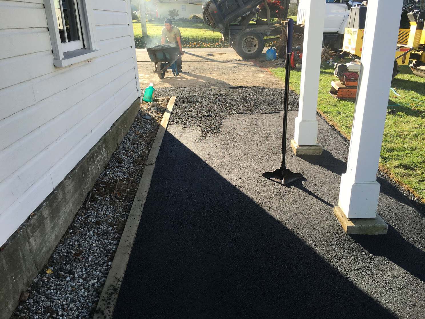 Crew member with a wheelbarrow of asphalt working on a narrow driveway beside a white house, with a dump truck and compactor behind