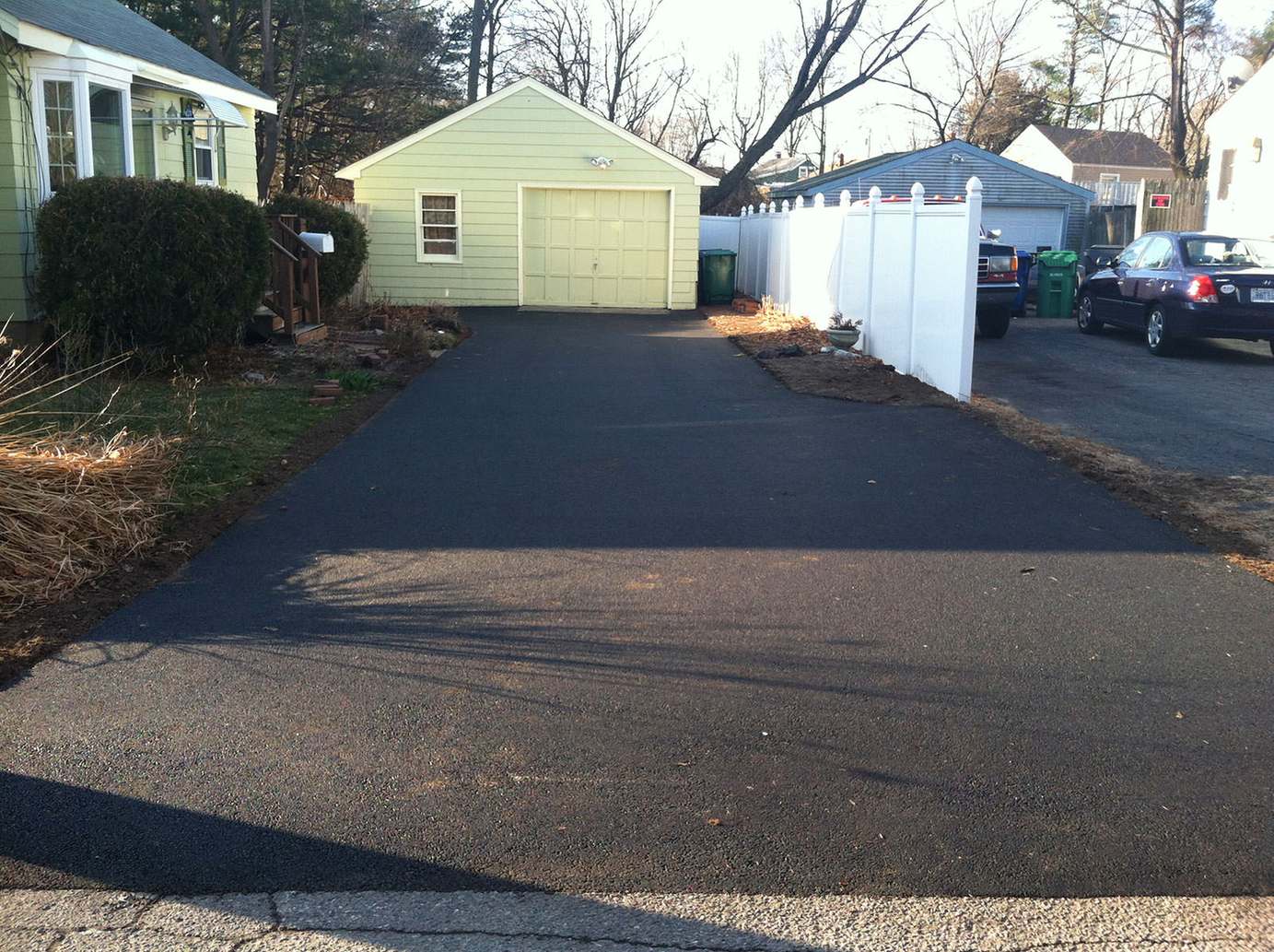 Newly paved residential asphalt driveway leading to a detached yellow garage beside a white vinyl fence