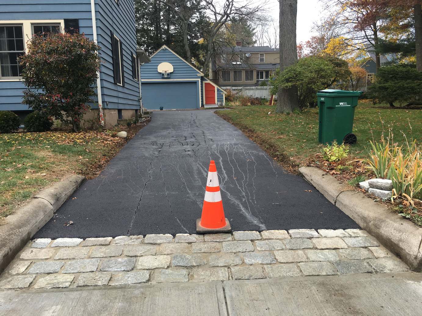 Newly overlaid asphalt driveway meeting a granite cobblestone apron at the street, with a traffic cone and a blue house in the background