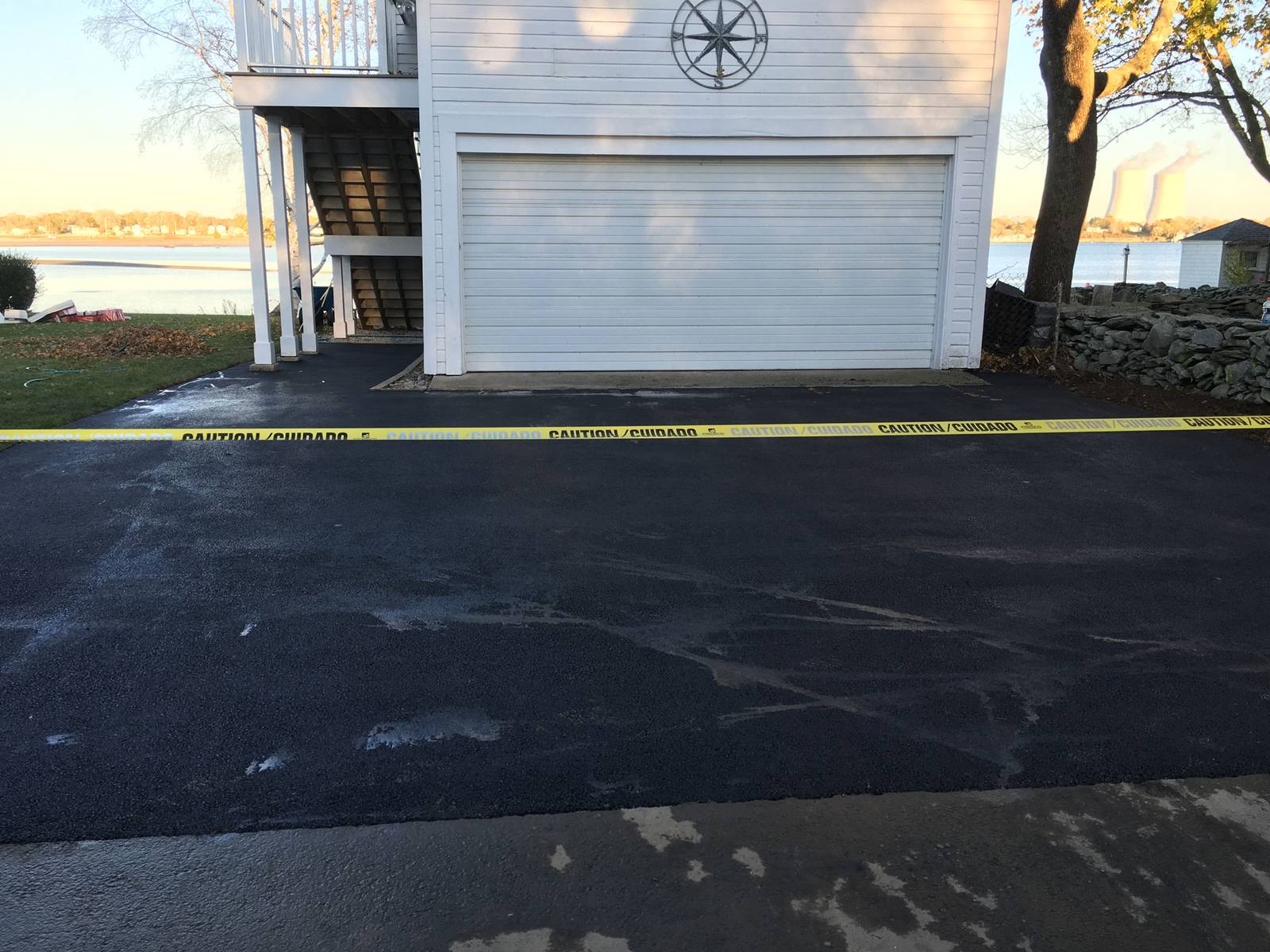 Fresh blacktop overlay in front of a white two-car garage near the water, with yellow caution tape across the new asphalt