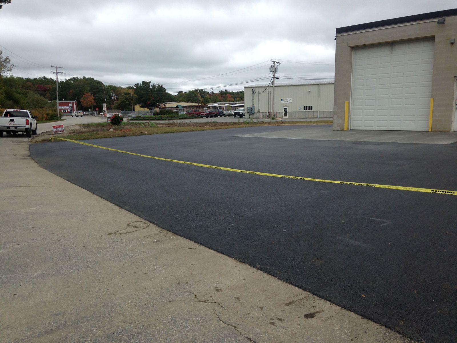 Freshly paved asphalt lot beside a commercial building with a large overhead door, with yellow caution tape marking the new pavement