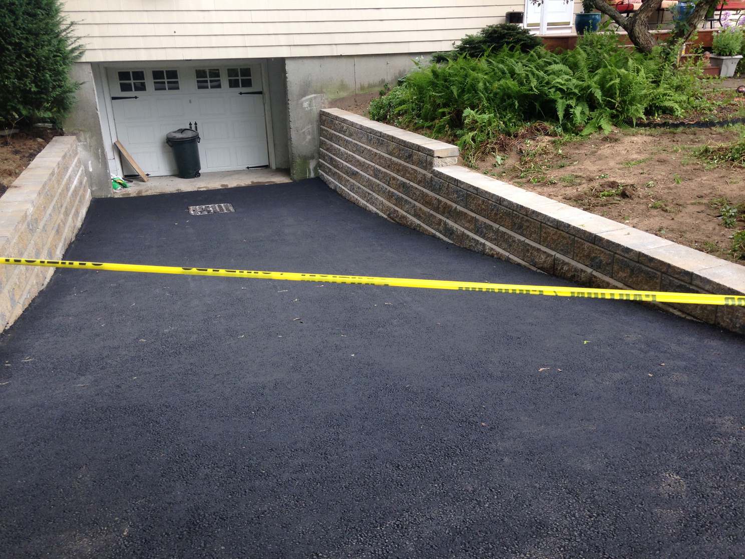 Freshly paved sloped asphalt driveway down to a basement garage with a block retaining wall and yellow caution tape