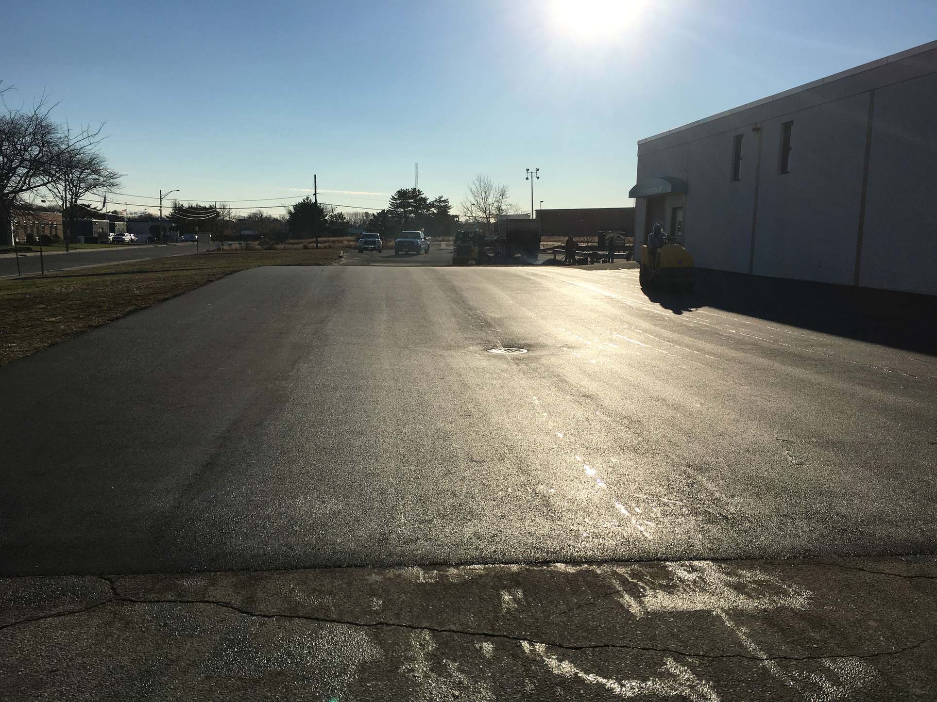 Large commercial parking area with freshly paved asphalt in low sun, with a paving crew and equipment in the background