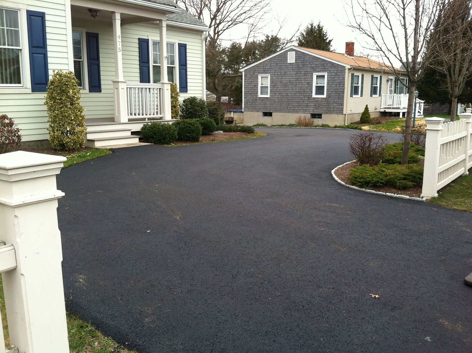 Newly paved curved asphalt driveway in front of a New England style house with blue shutters and a white picket fence
