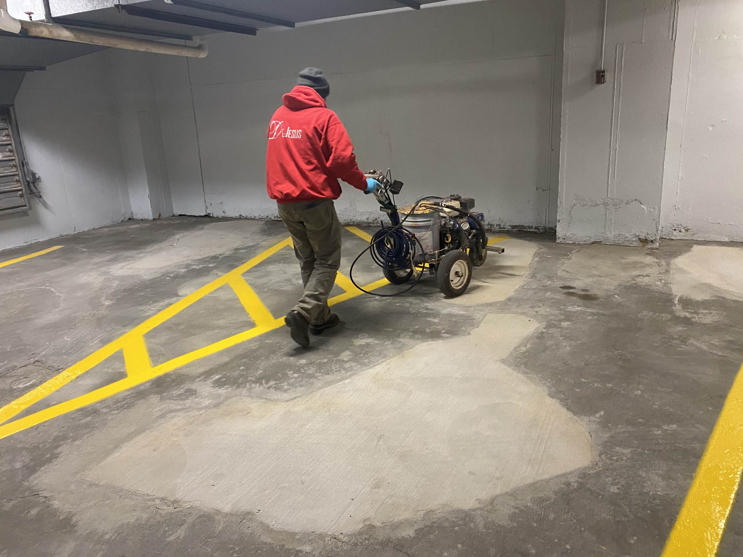 Crew member in a red DeJesus sweatshirt pushing a line striping machine across a concrete parking garage floor with fresh yellow lines