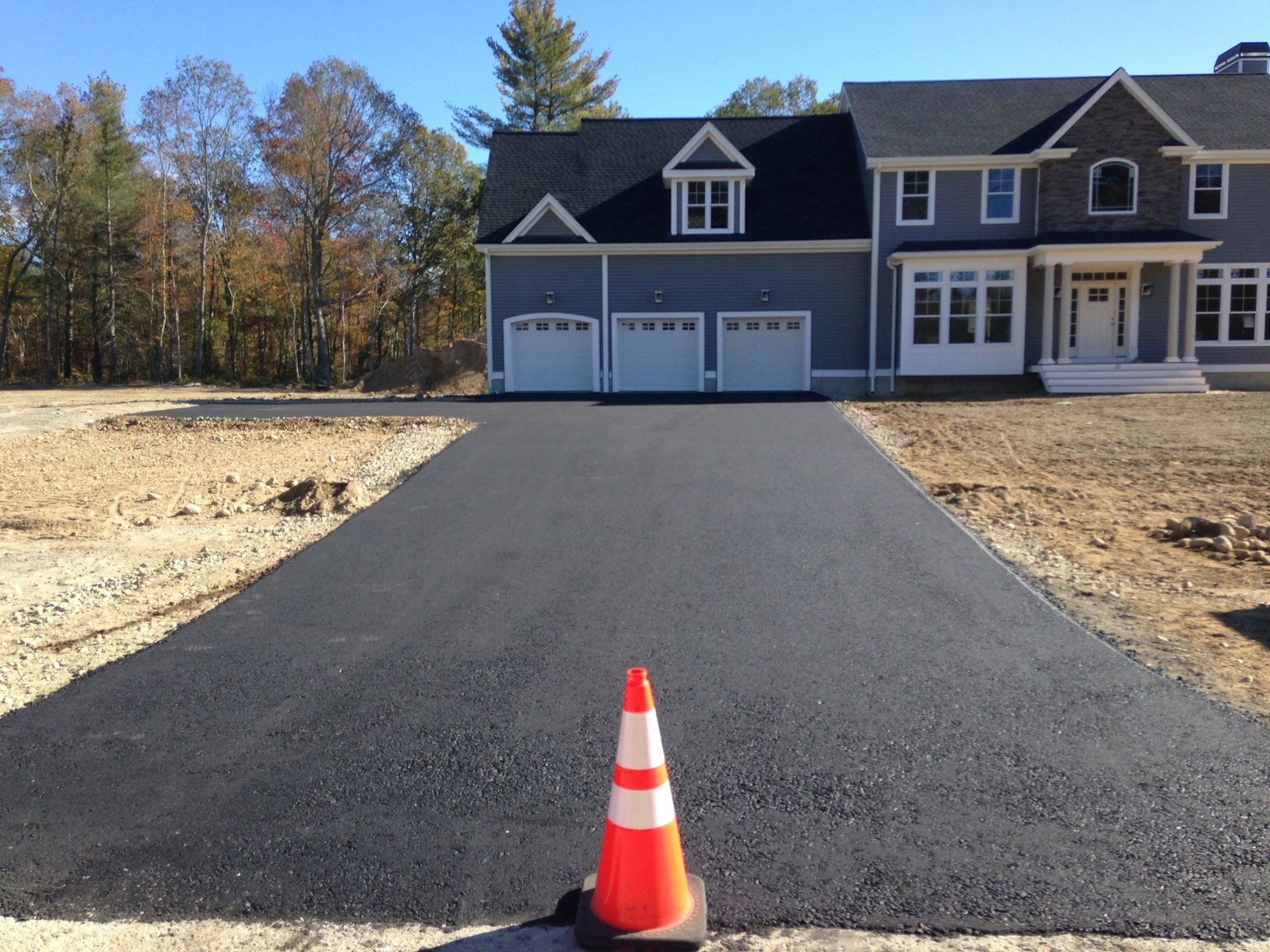 Freshly paved black asphalt driveway leading to a large gray home with a three-car garage, with a traffic cone in the foreground