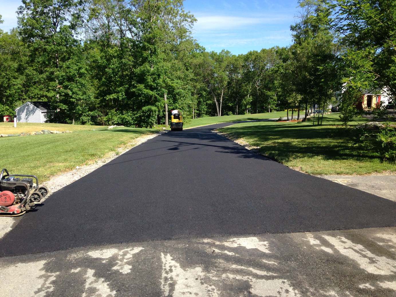 Newly paved private road curving between lawns and trees, with a roller and plate compactor at the edge of the new asphalt